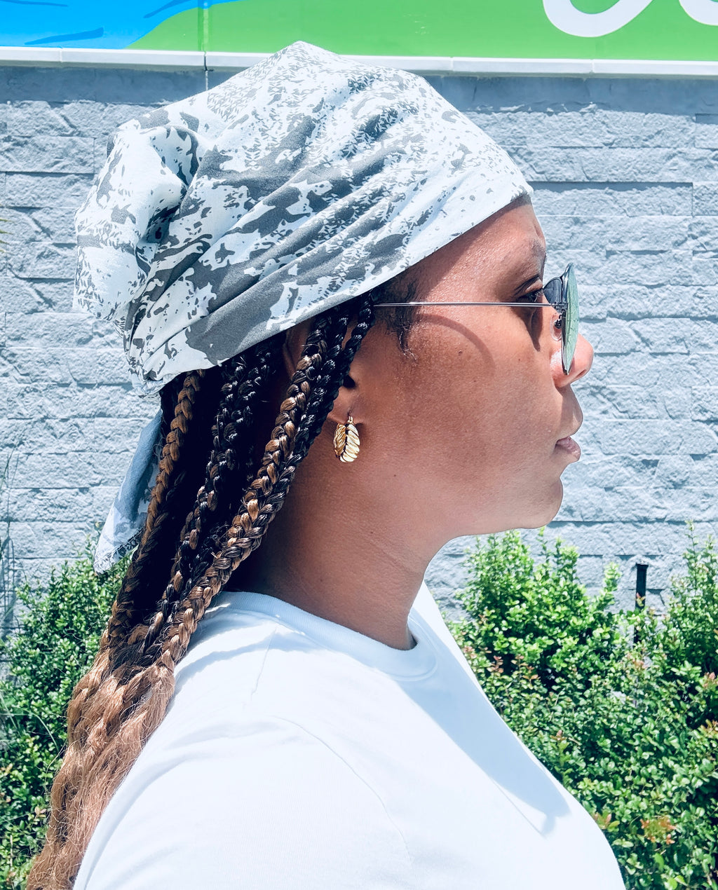 A woman in profile wearing a white and gray patterned headwrap, gold hoop earrings, and glasses, standing outdoors against a gray brick wall and green foliage.