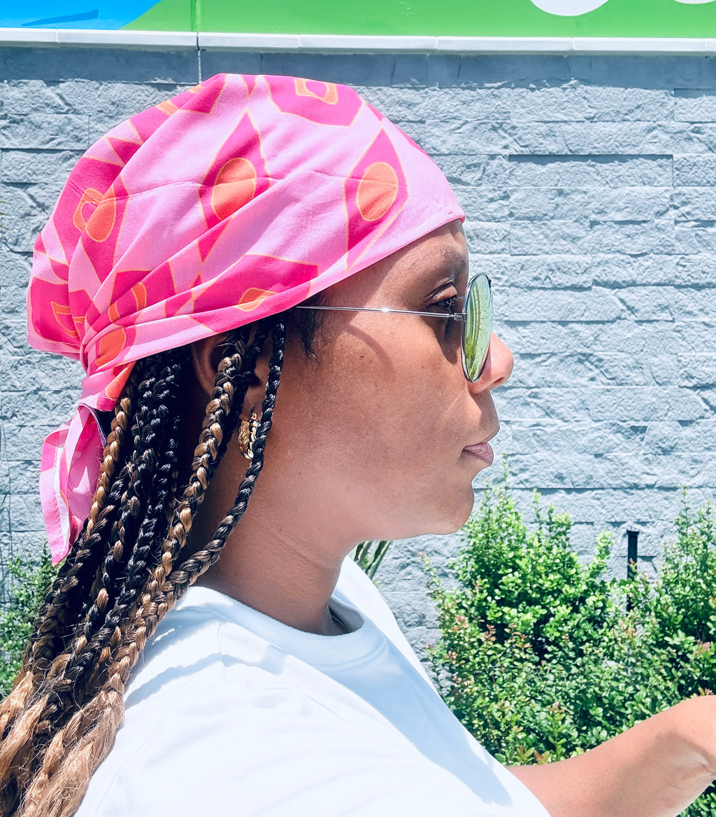 A woman in profile wearing a vibrant pink patterned headwrap and sunglasses, with braided hair and a white t-shirt, set against a gray brick wall background.