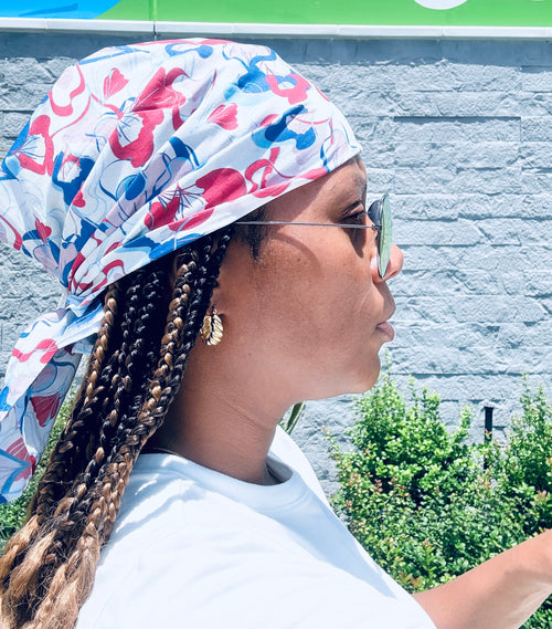 A woman in profile wearing a white headwrap with a blush pink, red, and blue floral pattern, styled with braided hair, sunglasses, and a white t-shirt against a gray brick wall background.