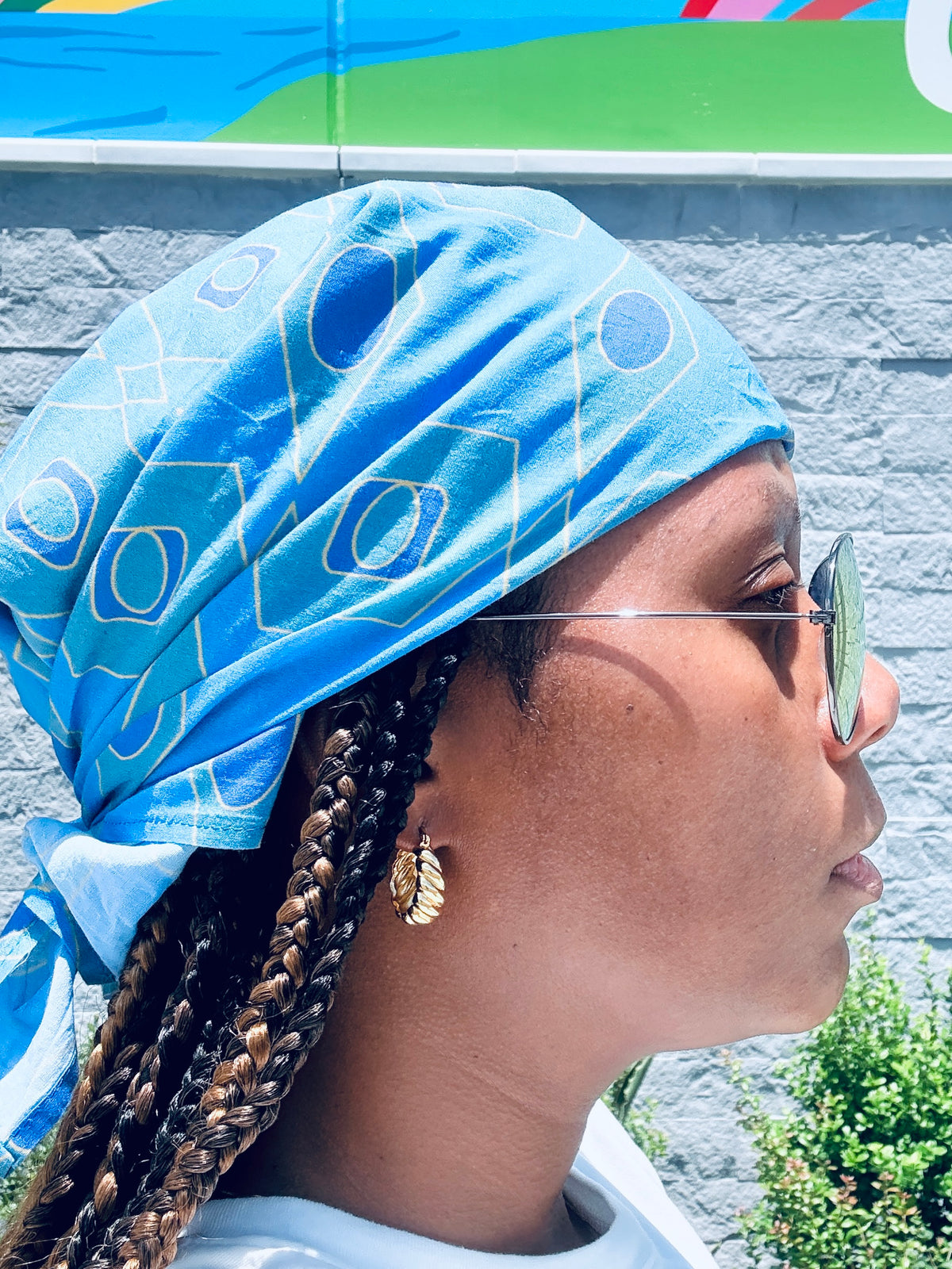 A close-up side profile of a person wearing a bright blue patterned headwrap, gold hoop earrings, and sunglasses, standing outdoors against a gray wall.