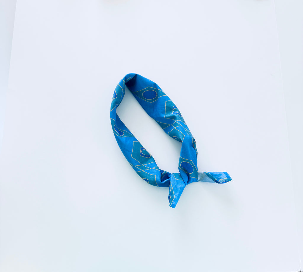 A blue patterned fabric headband with a geometric design, knotted at one end, displayed on a clean white background.
