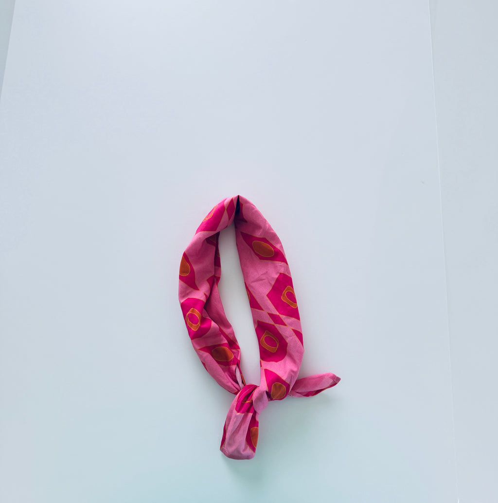 A pink fabric headband with a bold floral pattern in red, orange, and green, tied in a knot at the bottom, displayed on a clean white background.